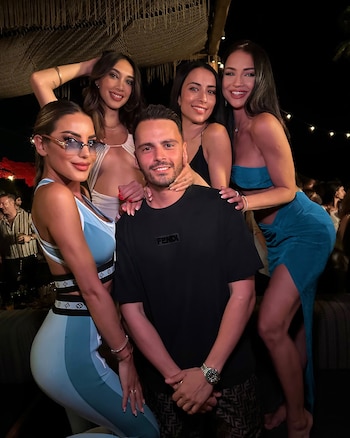 Un hombre y cuatro mujeres sonríen mientras posan juntos en un ambiente nocturno. El hombre, en el centro, viste una camiseta negra. Detrás, luces y techo tejido