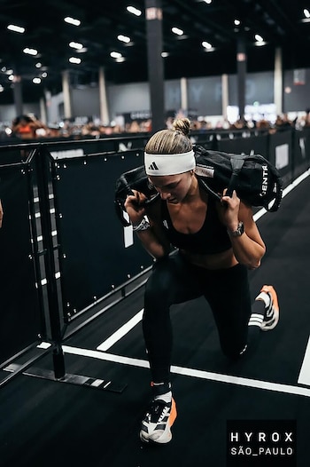 Camila Della Peña, vestida con un top blanco y pantalones cortos grises, se estira en un gimnasio. Se observan zapatillas, calcetines Adidas y una pelota medicinal en el suelo