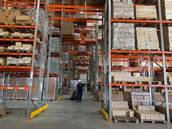 Almacén industrial con pasillos largos, estanterías altas cargadas de palets y cajas. Un operario con casco blanco y uniforme oscuro manipula una transpaleta en el pasillo