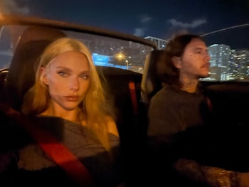 Una mujer rubia mira a la cámara y un hombre conduce un automóvil convertible de noche ante los edificios iluminados de una ciudad