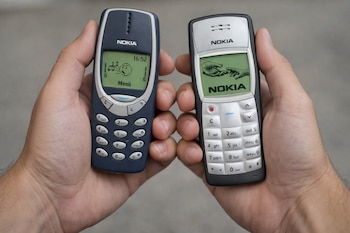 Primer plano de dos manos sosteniendo teléfonos móviles Nokia clásicos: un Nokia 3310 azul en la mano izquierda y un Nokia 1100 plateado en la derecha.
