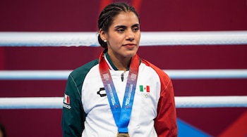 Mujer con chaqueta deportiva de México y una medalla al cuello frente a las cuerdas de un cuadrilátero.