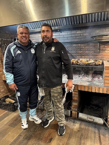 Foto de dos hombres sonrientes frente a una parrilla de ladrillos con un gran corte de carne asándose sobre brasas encendidas y extractor metálico