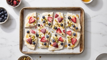 Trozos de yogur helado con fresas, arándanos, pistachos y miel sobre papel vegetal en una bandeja metálica rodeada de cuencos.