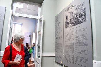 Una mujer con cabello canoso y gafas observa paneles de exposición. Un panel principal exhibe una ilustración histórica y el título 'Barranco, Ciudad Heroica'