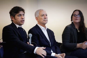 Axel Kicillof, un hombre de cabello oscuro y camisa blanca, habla a un micrófono gris mientras está sentado junto a un hombre y una mujer