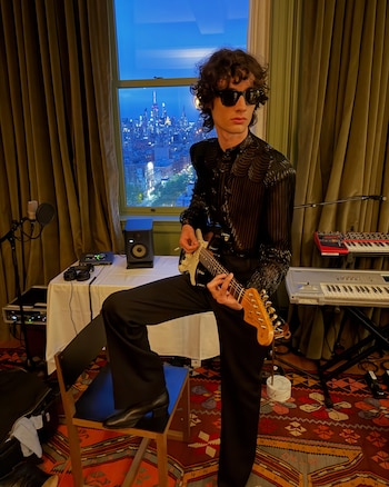 Joven con gafas de sol tocando guitarra eléctrica, sentado en un taburete frente a una ventana con vista a un skyline nocturno de ciudad y equipos de audio