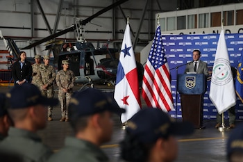 Un hombre habla desde un podio junto a las banderas de Panamá y Estados Unidos, con militares y un helicóptero en un hangar de fondo