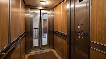 Interior de un ascensor con paneles de madera, botonera metálica, alfombra café y puertas con ventanas de vidrio.