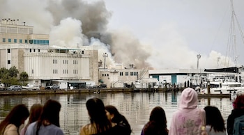 Vista de un edificio blanco en llamas con humo denso elevándose, con bomberos combatiendo el fuego. Personas observan desde una orilla con botes anclados