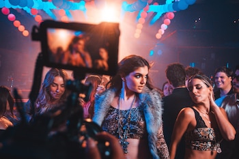 Miranda Capurro con top estampado animal print y chaleco de piel sintética, junto a varias personas, es filmada por una cámara en un club con luces de colores