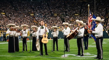 Integrantes de un mariachi tocan guitarras, violines y trompetas en la cancha de un estadio repleto de público.
