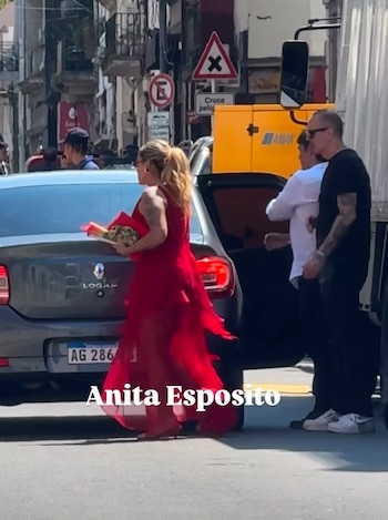 Una mujer con un vestido rojo largo camina en la calle junto a un automóvil gris con la puerta abierta y dos hombres