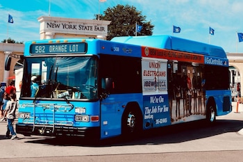 Autobús azul estacionado frente a una entrada que dice Gran Feria Estatal de Nueva York, con personas, banderas y árboles