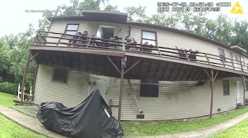 Una casa de dos pisos con terraza de madera semiderruida. Varios agentes de seguridad en la terraza. Una motocicleta cubierta con lona. Texto: US Marshals Service