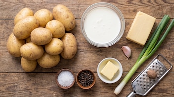 Papas, crema en bol de vidrio, bloque de queso, cebolla de verdeo, ajo, manteca en plato, sal, pimienta, rallador con nuez moscada en tabla de madera.
