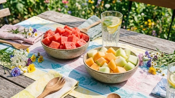 Una mesa de madera con dos cuencos de sandía y melón en cubos, un vaso de agua con limón y menta, cubiertos de madera, servilletas y flores.
