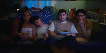 Cuatro jóvenes sentados en un sofá oscuro, tres comen de cuencos y uno tiene la mano en la cara; al fondo, mantas y almohadas