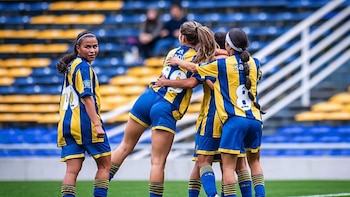 Cuatro mujeres futbolistas con uniformes a rayas azules y amarillas. Tres de ellas se abrazan mientras una mira a la cámara. Fondo de tribunas