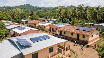 Vista aérea de un pueblo en El Salvador con casas de techos de lámina y teja, muchas con paneles solares. Se ven personas, una camioneta y caminos de tierra.