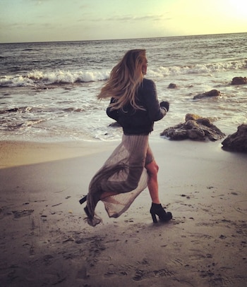Mujer rubia de espaldas, con chaqueta oscura y falda larga vaporosa, corriendo en la orilla de una playa rocosa con el mar y un sol brillante