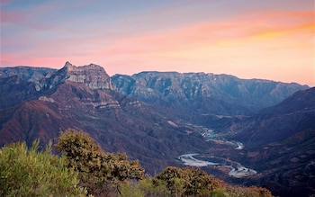 Montañas escarpadas cubiertas de vegetación, un río serpenteando en un valle profundo, arbustos en primer plano y un cielo con tonos rosados y anaranjados