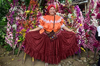 Mujer de mediana edad con blusa naranja y falda roja posa frente a una gran variedad de flores coloridas como malvas reales y delphinium