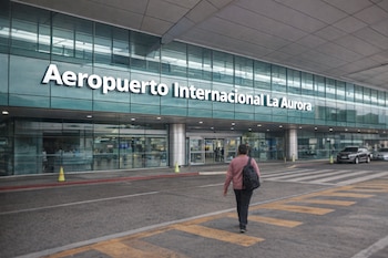 Vista frontal del Aeropuerto Internacional La Aurora con su nombre en la fachada de cristal, una persona caminando y vehículos estacionados cerca de la entrada.