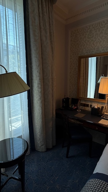 Interior de una habitación de hotel con ventana, cortinas, una lámpara, un escritorio con silla, un espejo de pared con marco dorado, alfombra y parte de una cama