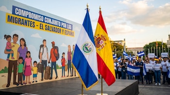 Dos banderas, una de El Salvador a la izquierda y una de España a la derecha, en primer plano. Detrás, un cartel ilustrado sobre el compromiso por los migrantes.