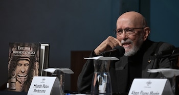 Un hombre calvo con barba y gafas habla en un podio con dos micrófonos, dos carteles de identificación, un libro y dos vasos con agua