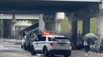 Un SUV policial blanco con luces rojas y azules intermitentes en una calle mojada, bajo una estructura elevada de hormigón. Se ven otros coches y una persona con paraguas