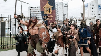 Grupo de personas con vestimenta y cascos medievales que portan espadas, hachas, escudos de madera y un estandarte en una calle