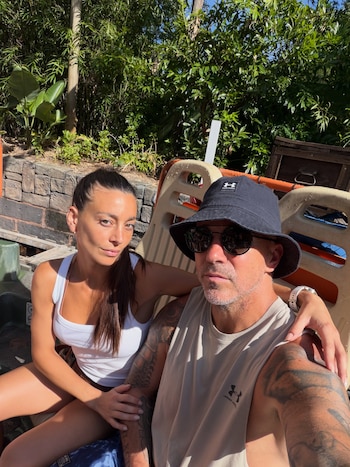 Una mujer con camiseta de tirantes blanca y un hombre con sombrero negro, gafas de sol y tatuajes en el brazo