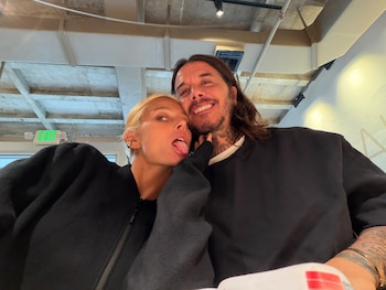 Una mujer rubia saca la lengua junto a un hombre sonriente con tatuajes en un espacio interior con techo industrial