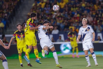 Dos jugadores de fútbol saltan para cabecear un balón en el aire; uno viste uniforme amarillo y otro uniforme blanco, con más jugadores y espectadores en un campo de juego