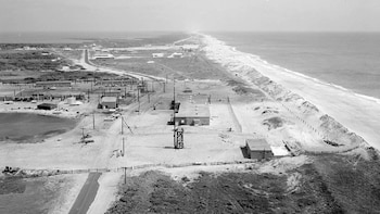 Vista aérea en blanco y negro de una zona costera con edificios, carreteras, una laguna, y una extensa playa de arena con dunas