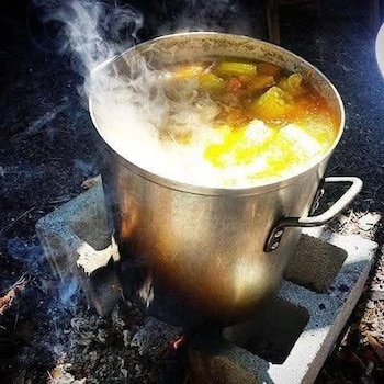 Primer plano de una olla grande de metal hirviendo con un líquido amarillento y trozos de maíz u otros vegetales, sobre un fuego abierto