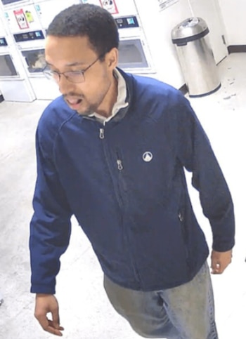 Hombre con gafas, chaqueta azul y jeans, mirando hacia abajo, captado por cámara de seguridad en una lavandería con máquinas blancas al fondo