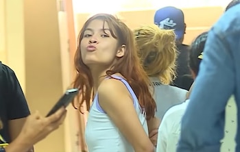 Una mujer joven de cabello castaño rojizo y top blanco frunce los labios en un gesto de beso mientras mira a la cámara, rodeada de personas