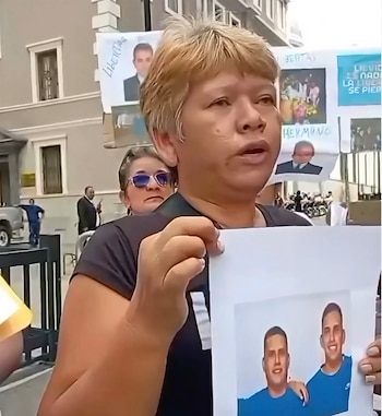 Una mujer de cabello rubio, con camiseta negra, sostiene una foto de dos hombres sonrientes; en el fondo, otras personas y pancartas se ven frente a un edificio