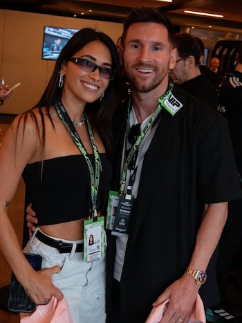 Antonela Roccuzzo con top negro y gafas de sol junto a Lionel Messi con chaqueta negra y reloj dorado, ambos sonriendo y mostrando pases VIP en un evento cubierto