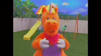 Un personaje disfrazado de color naranja con cuerno y pañuelo rosado frente a un tobogán amarillo y columpios.