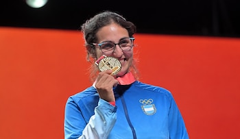 Una mujer con gafas y chaqueta deportiva azul sostiene una medalla dorada junto a su rostro sobre un fondo rojo
