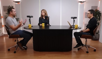 Tres personas sentadas alrededor de una mesa negra con micrófonos, tazas amarillas y un ordenador portátil. Paredes blancas, piso de madera, plantas