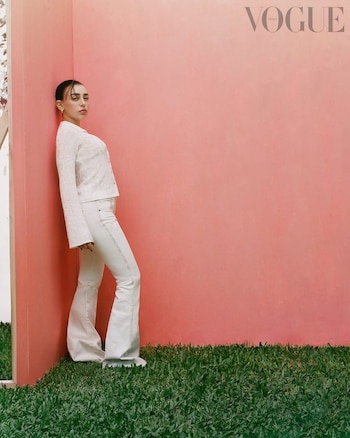 Nicki Nicole de cuerpo entero, con atuendo blanco texturizado y pantalones acampanados, apoyada en una pared rosa pálido con césped verde en el suelo