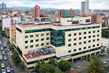 Edificio con paredes de piedra clara y ventanas verdes, con el letrero amarillo y blanco 'salup Clínica Infantil'. Calles con autos, árboles y edificios urbanos.