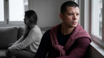 Hombre de cabello corto y suéter rojo atado al cuello, mirando por una ventana, con una mujer desenfocada y en escala de grises de espaldas al fondo.