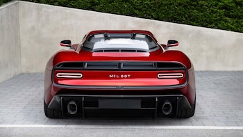 Vista trasera de un automóvil deportivo rojo con luces horizontales, logotipo MCL 6GT, dos tubos de escape y difusor. Fondo de pared clara y vegetación