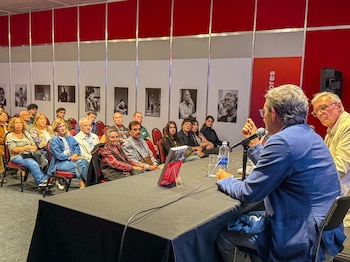 Dos hombres sentados en una mesa con micrófonos hablando a una audiencia sentada en sillas rojas. Un libro y una botella de agua están sobre la mesa. Detrás de la audiencia hay fotografías en blanco y negro enmarcadas